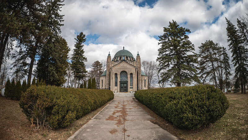 Classical chapel with green dome centered between symmetrical hedges and tree-lined pathway.