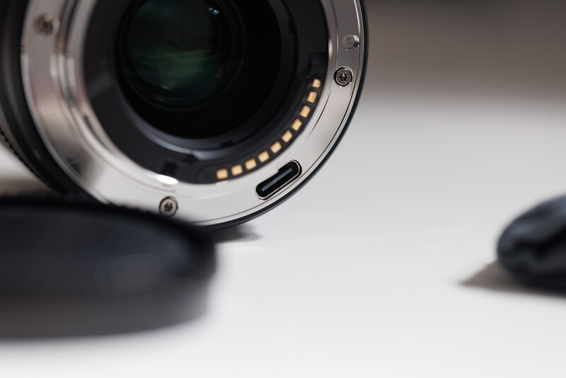 Close-up of a camera lens mount showing electronic contacts and mechanical bayonet ring against a neutral background.