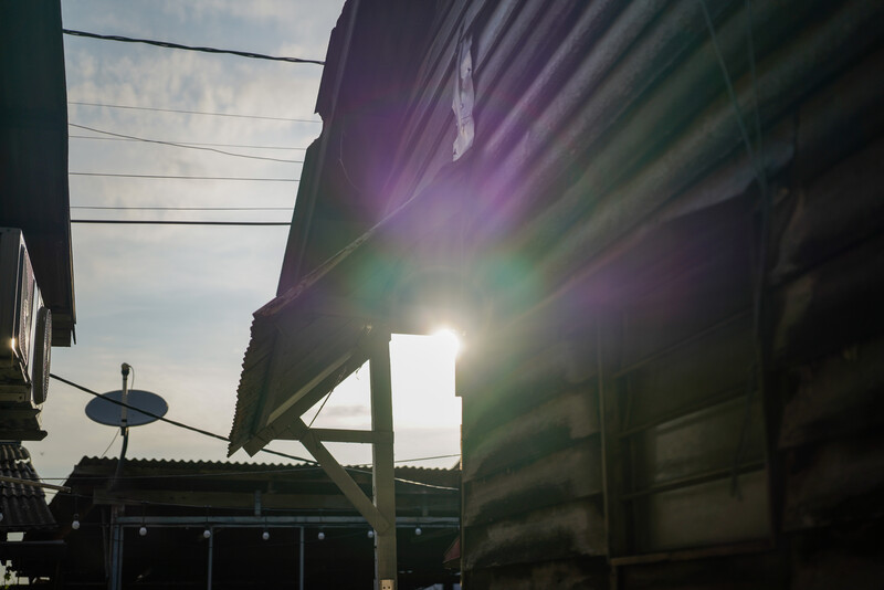 Bright sun flare streaming through industrial metal structures and power lines, creating dramatic lens flare and purple-green color casts.