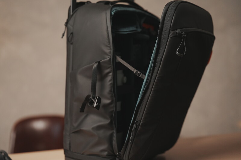 Black camera backpack displayed open, showing internal compartments and organization with a wooden chair visible in soft, warm background lighting.