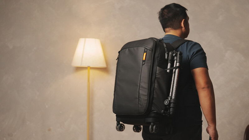Man wearing dark backpack and blue shirt stands facing wall with illuminated floor lamp.
