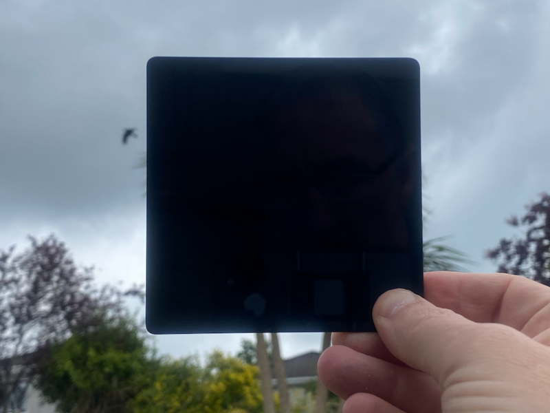 Hand holding a dark neutral density filter against a cloudy sky with trees in the background.