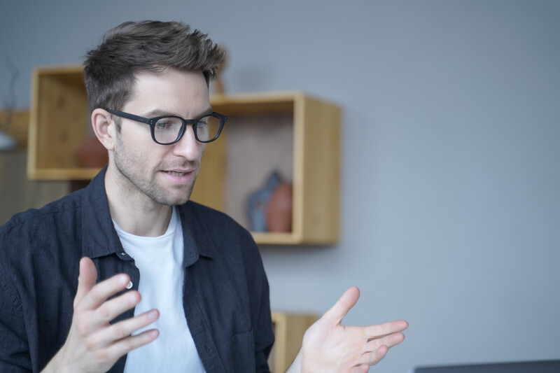 Man wearing glasses and dark jacket gestures while speaking indoors.