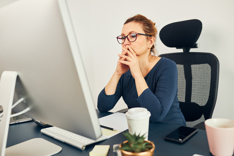 Woman at desk with laptop, wearing glasses and dark cardigan, chin resting on hand in contemplative pose.