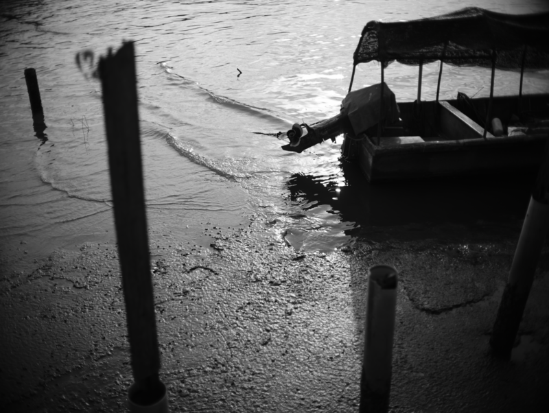 Silhouetted figure sitting in a wooden boat on shallow water, photographed in high-contrast black and white.