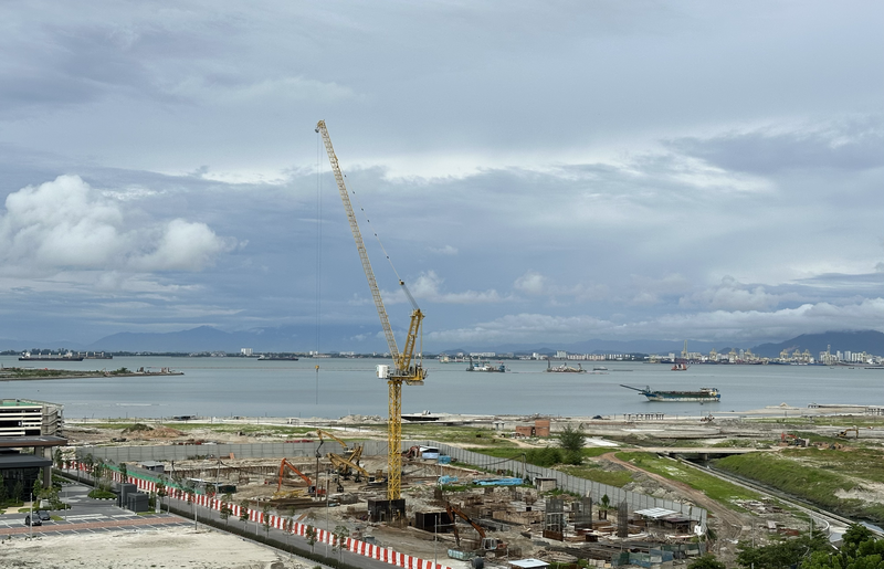Construction site with yellow crane extending over waterfront industrial area under dramatic cloudy sky.