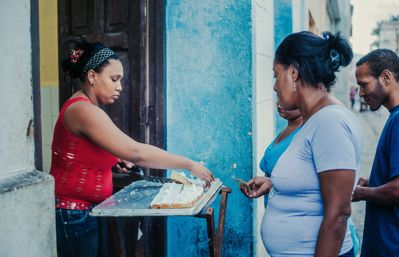 Woman in red tank top serving food from a tray to customers on a street.