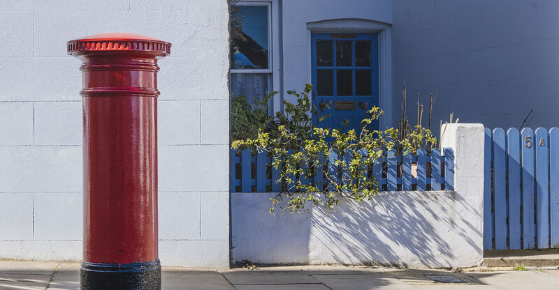 Red British postal box on white-walled street with blue door and gate in background.