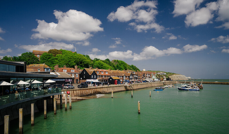 Waterfront village with brick buildings, wooden pier, and turquoise harbor water under blue sky with white clouds.