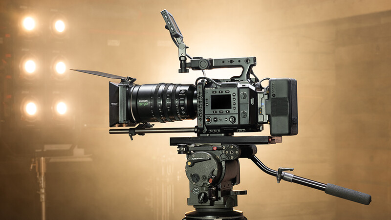 Professional cinema camera with prime lens, matte box, and microphone mounted on fluid head tripod under studio lights.