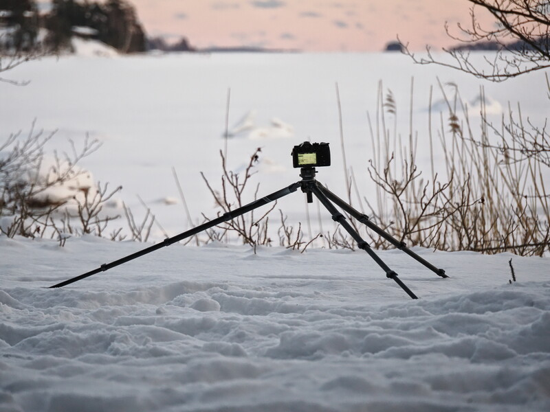 Action camera mounted on tripod legs in snowy landscape with frozen lake and bare vegetation in background.