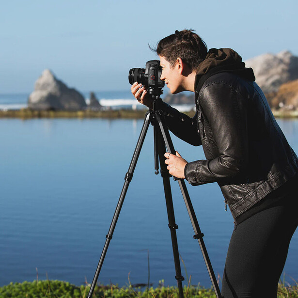 Photographer using a tripod-mounted DSLR camera to capture a scenic landscape with calm water and distant hills.
