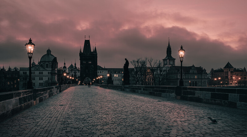 Historic stone bridge at dusk with Gothic architecture and lit street lamps under dramatic purple-pink sky.