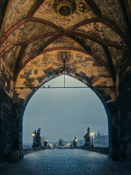 Gothic archway framing a distant bridge and cathedral spires across a cobblestone plaza.