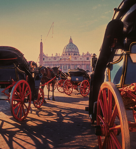Horse-drawn carriages with red wheels lined up in a plaza, a domed basilica in the background at golden hour.