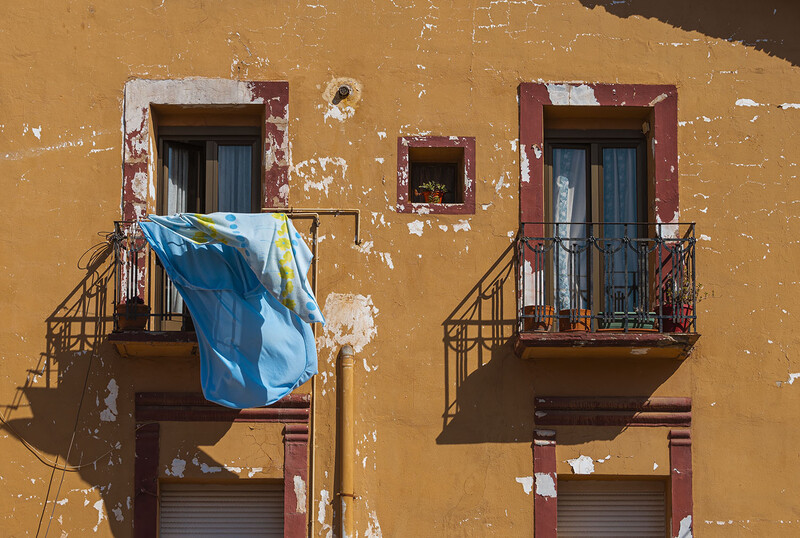 Weathered yellow building facade with two red-trimmed windows, blue fabric hanging from left balcony.