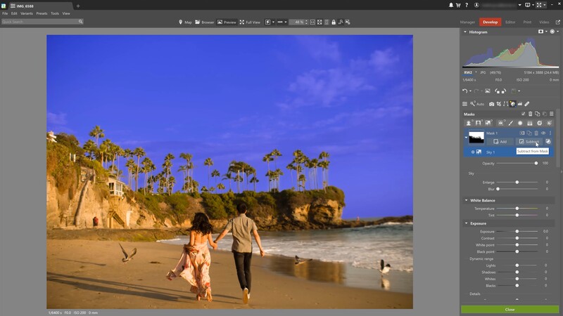 Two people walking along a sandy beach with palm trees and golden cliffs under a vibrant blue sky.