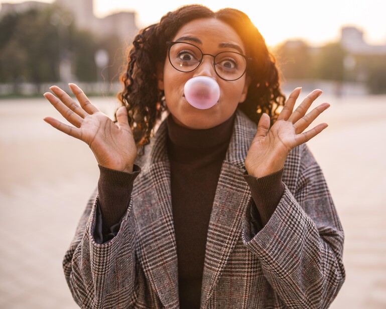 Person with glasses blowing a pink bubble gum bubble outdoors during golden hour.