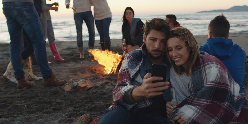 Couple taking a selfie at a beach bonfire gathering at dusk.