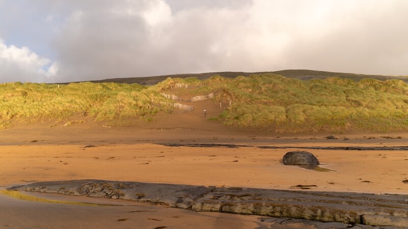 Sandy beach with exposed rock formations and vegetated cliffs under dramatic cloudy sky.