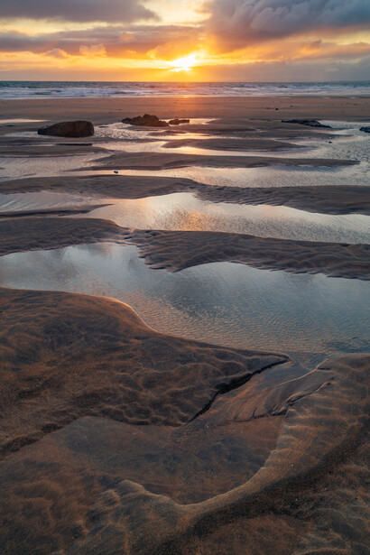 Tidal flats at sunset with shallow pools reflecting golden sky and dark sand formations.