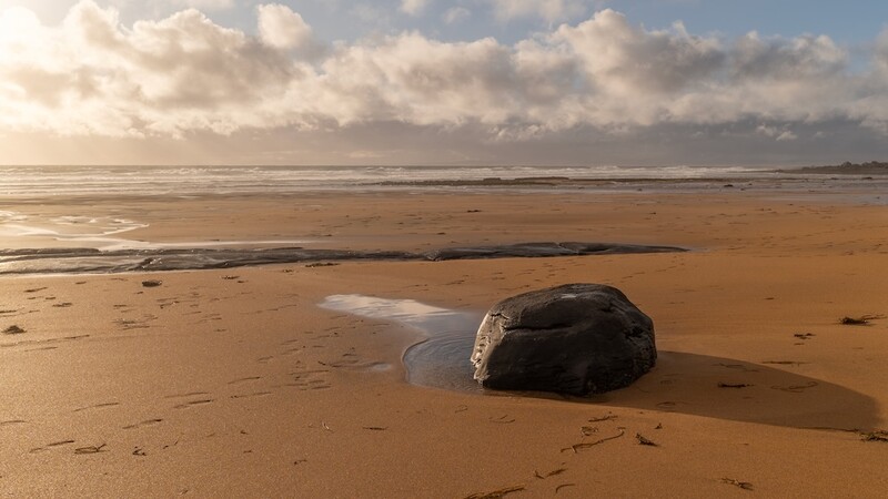 Dark rock partially submerged in shallow water on a wide sandy beach under cloudy sky.