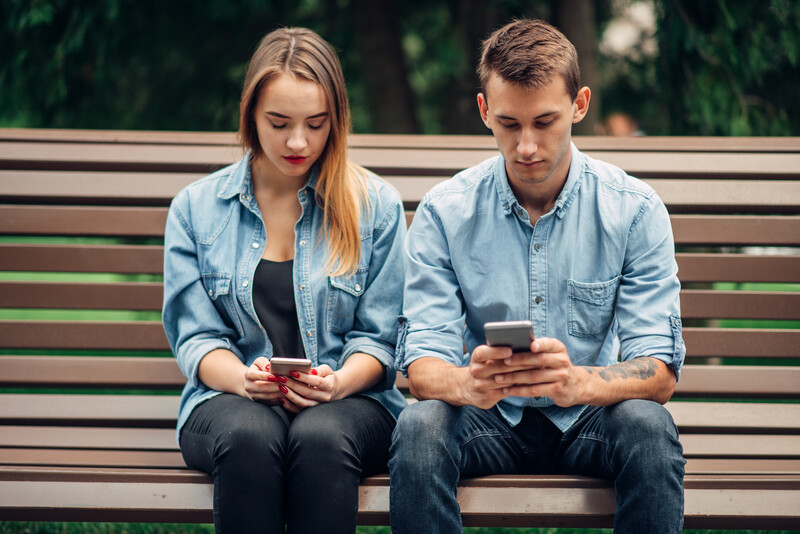Two young adults sitting on a bench, both focused on their smartphones.