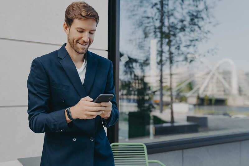 Smiling man in dark blazer looking at smartphone outdoors near modern architecture.