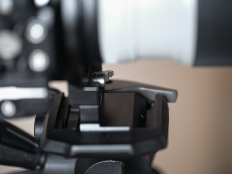 Close-up of camera iron sights mounted on a rifle, showing the rear peep sight in sharp focus with blurred equipment and workspace in background.