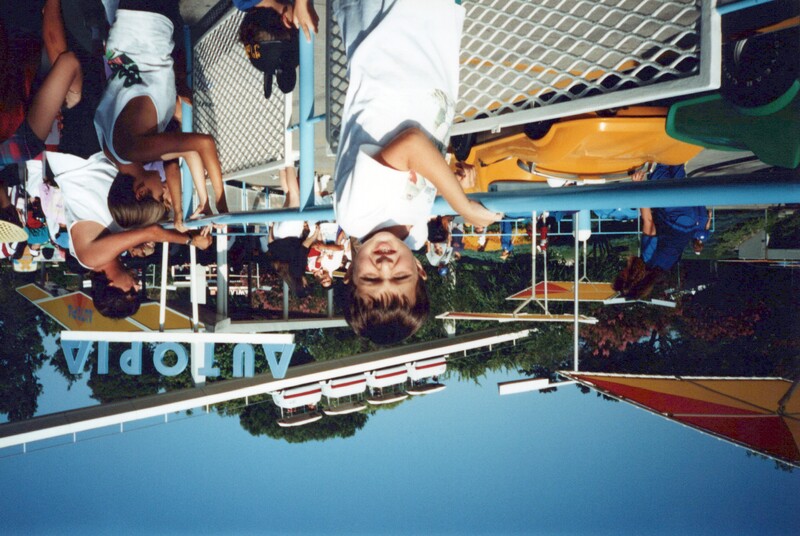 Young child in white shirt posing at an amusement park ride entrance on a sunny day.