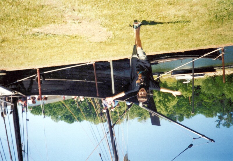 Person in black shirt striking an energetic pose on grass in front of a beached wooden sailing vessel.