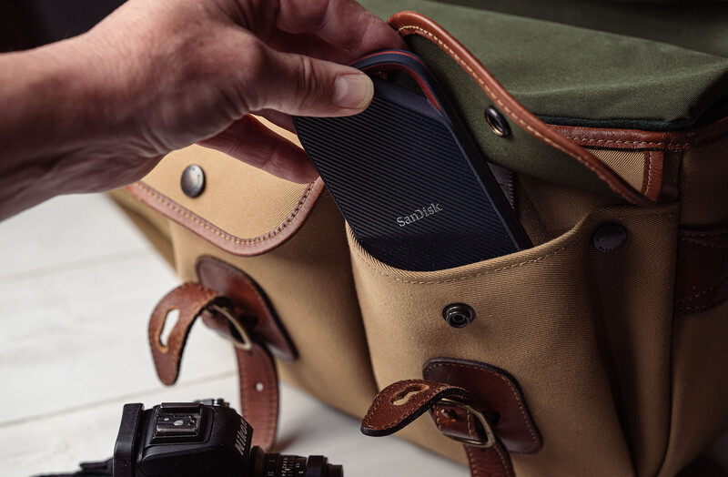 Tan canvas camera bag with brown leather accents holds a black external hard drive and other photography gear.