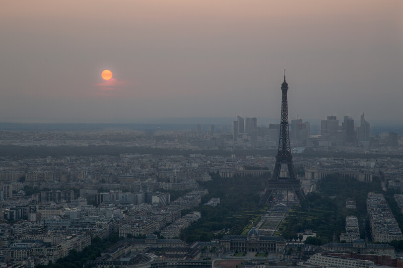 Eiffel Tower silhouette at dusk with orange sun and distant cityscape.