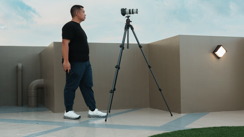 Photographer standing beside a tripod-mounted telephoto lens on a rooftop with geometric concrete structures.