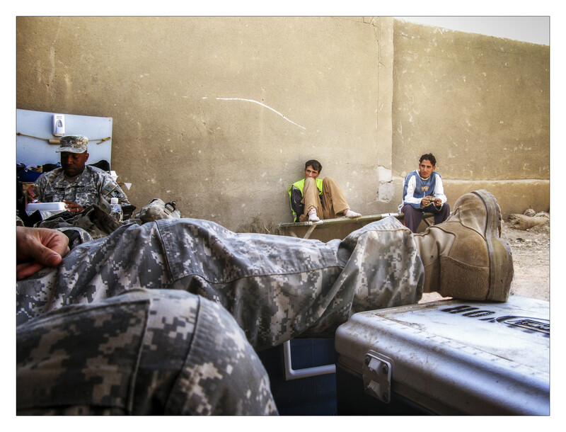 Two people sit against a weathered tan wall beside military-style camouflage clothing and worn luggage.