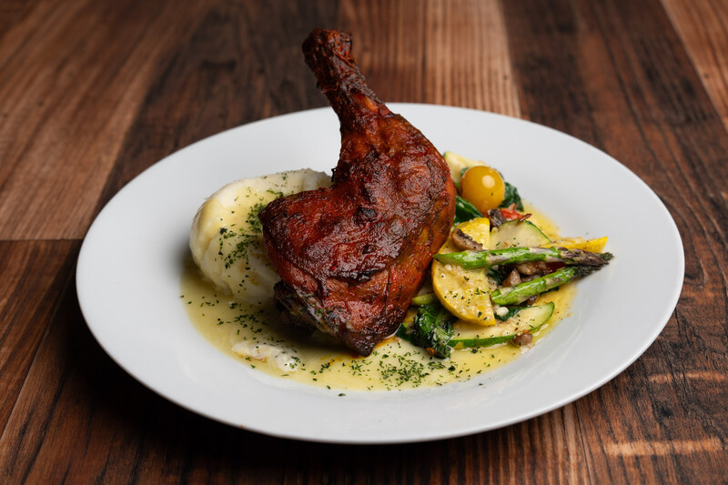 Roasted duck leg with potato puree and grilled vegetables on white plate with sauce drizzle.