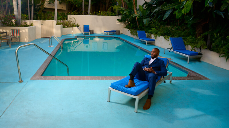Man in dark suit sitting on blue lounge chair beside empty turquoise swimming pool.