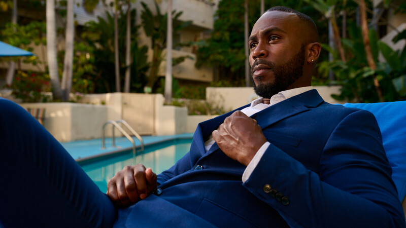 Man in navy blazer adjusting tie by poolside with palm trees in background.