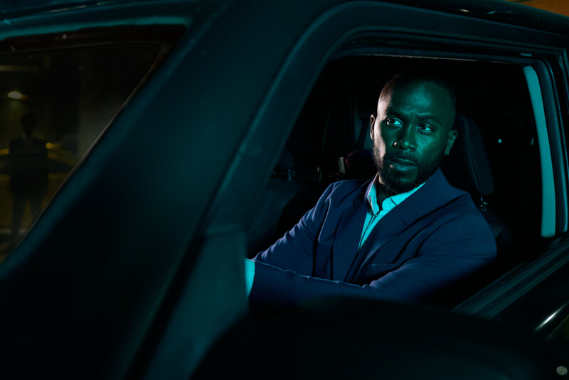 Man sitting in driver's seat of car at night, lit by cool teal and cyan interior lighting.