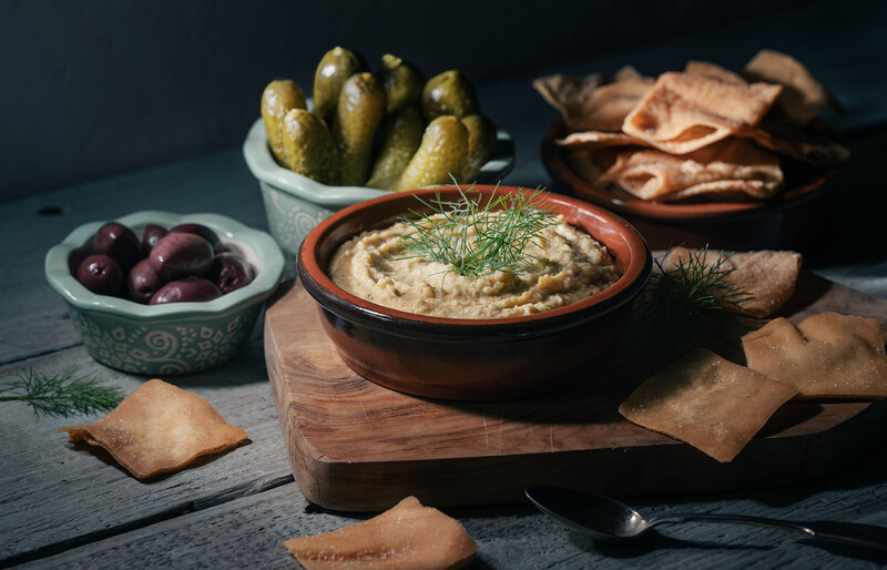 Hummus in a terra cotta bowl garnished with dill, surrounded by pickles, olives, and crackers on a dark surface.