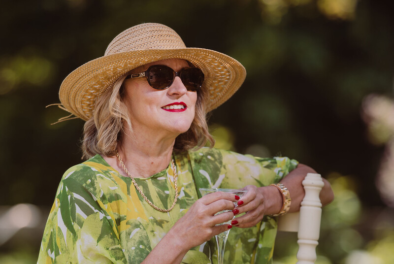 Woman wearing sunglasses and a straw hat, dressed in a green floral patterned shirt, posed outdoors in natural sunlight.