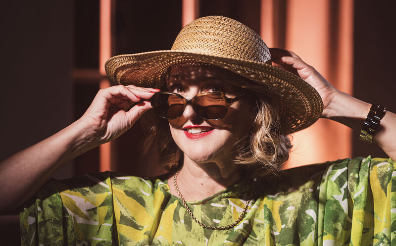Woman wearing a straw hat and sunglasses adjusts her frames while posing in warm, directional sunlight.