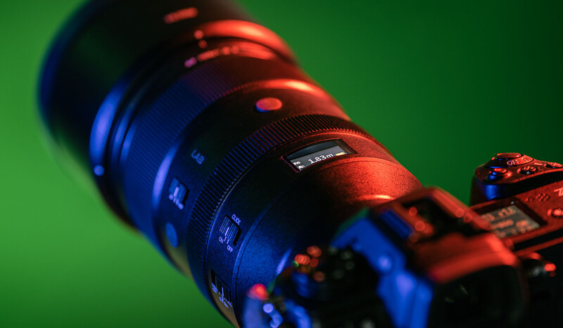Camera lens with focus ring and depth-of-field markings, lit with red and blue gels against green background.