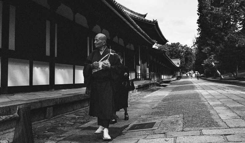 Elderly person in traditional dark formal wear standing in front of a Japanese temple building.