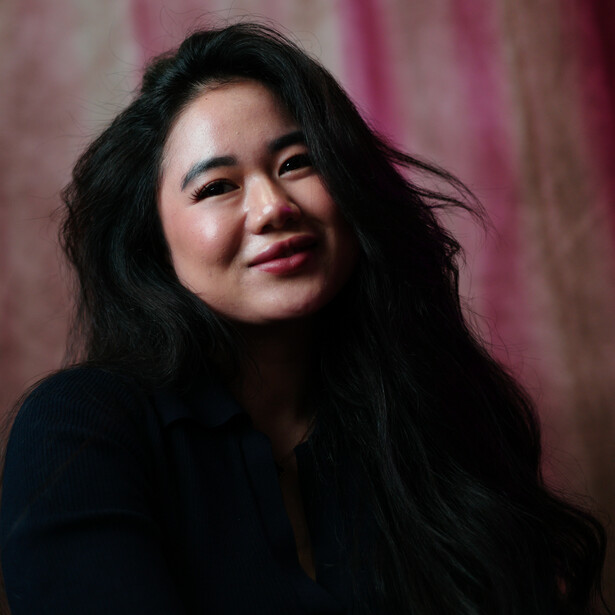 Woman with long dark hair smiling at camera against pink-toned backdrop.