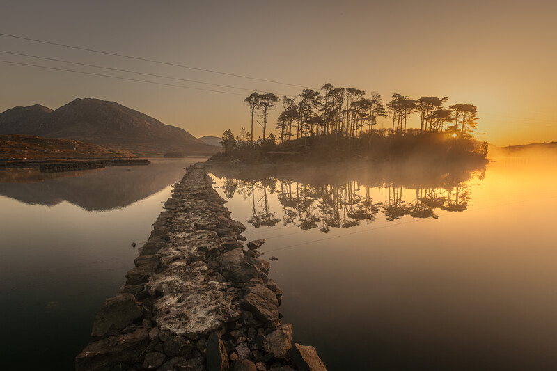 Rocky causeway extends into a misty lake at golden sunrise, with forested island and mountains reflected in still water.