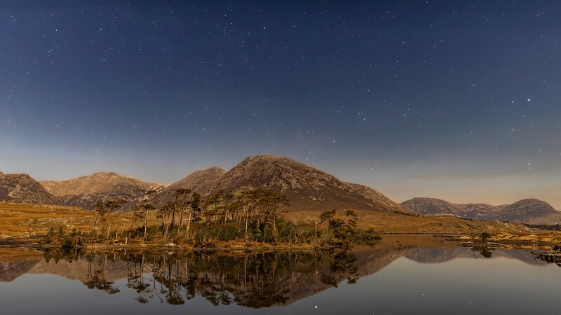 Mountain landscape with palm trees reflected in still water under a starry night sky.