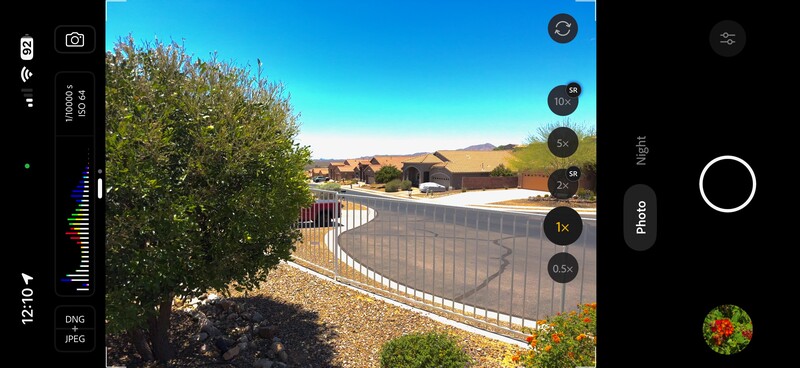 Mobile phone camera interface displaying a residential street with white fence and desert mountains in background.
