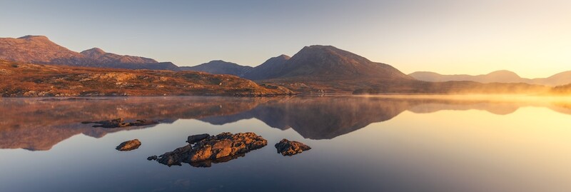 Mountain landscape reflected in still water at sunrise, with rocky outcrops in the foreground.