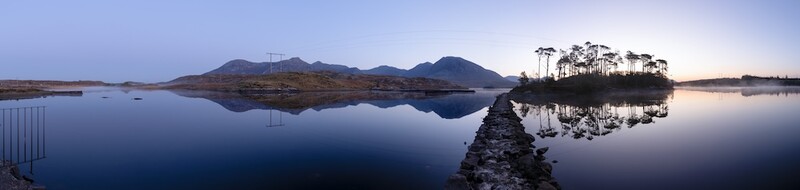 Still lake at dawn with rocky pier leading to forested island and mountain backdrop.